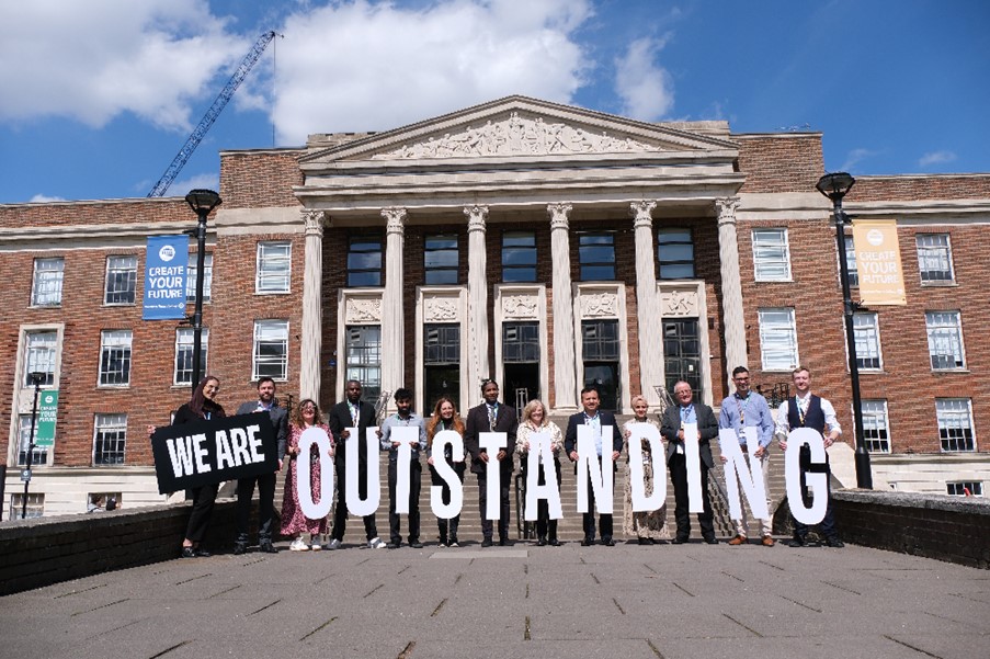 Students stand in front of the College holding letters that spell Outstanding
